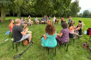 Venkovní bubenický jam v parku. Velká různorodá skupina lidí sedí v kruhu na trávníku a židličkách, každý s africkým bubnem. Účastníci všech věkových kategorií společně hrají v uvolněné atmosféře. Kolem jsou stromy a zelená louka, vytvářející příjemné přírodní prostředí pro spontánní hudební setkání.