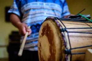 Detail afrického basového bubnu dunun s paličkou. V popředí je vidět napjatá kůže bubnu s charakteristickými tmavými skvrnami a barevnými provázky po obvodu. Bubeník v modře vzorované košili drží dřevěnou paličku, připraven k úderu. Záběr zachycuje moment těsně před zahráním, zdůrazňující texturu a konstrukci tradičního afrického nástroje.